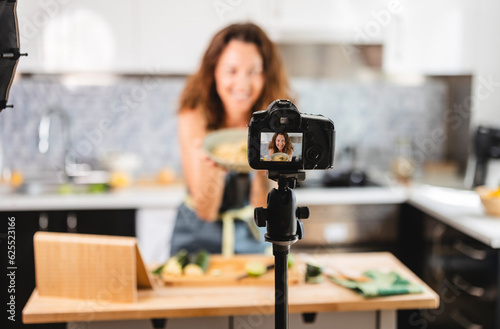 Mature woman talks to a digital camera on a tripod shows the ingredient while vlogging about making food recipes in the kitchen at home, social media.