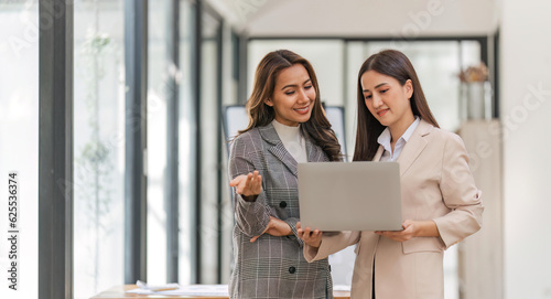 Two happy professional coworkers discussing plan at work. explaining Asian colleague financial project on laptop in office