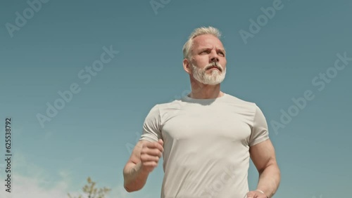 Low angle slow motion shot of athletic senior sportsman running outdoors on sunny summer morning with cloudless sky