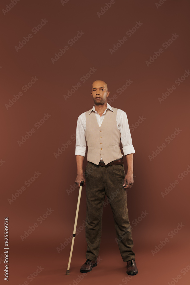 Fototapeta premium real people, bold african american man with myasthenia gravis syndrome standing with walking cane on brown background, standing and looking at camera, diversity and inclusion, physical impairment