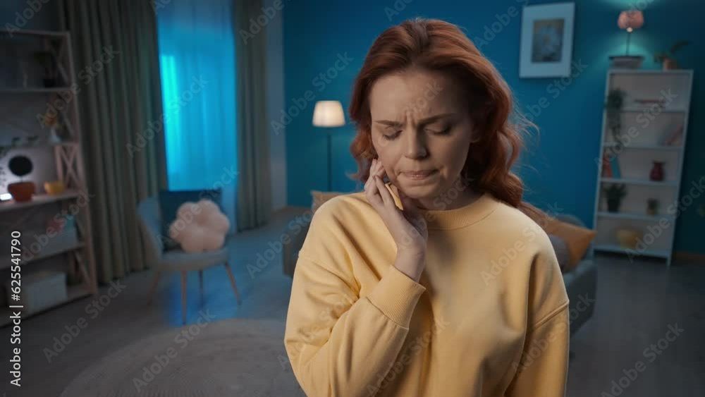 A young woman stands in the living room and holds her cheek close up. A