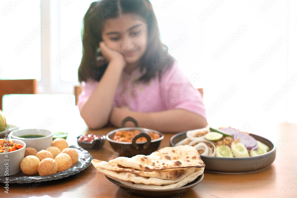 Indian child girl sitting at the dining table looking at traditional Indian food on the table and feeling bored, selective focus Indian child girl sitting at the dining table looking at traditional Indian food on the table and feeling bored, selective focus
