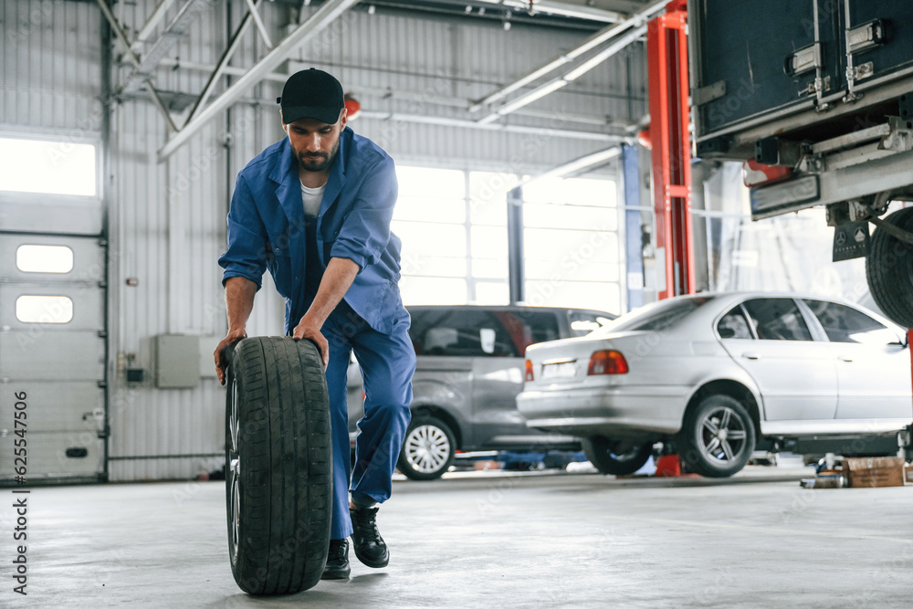 Walking forward with new tire. Auto mechanic working in garage. Repair service Walking forward with new tire. Auto mechanic working in garage. Repair service