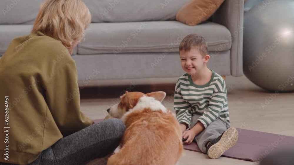 Cheerful 6 year old boy with down syndrome sitting on rug in bright living room playing with his mum and cute corgi dog