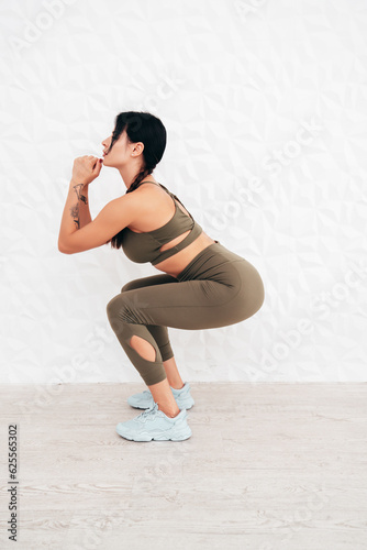 Portrait of fitness woman in sports clothing. Sexy young beautiful model with perfect body wearing sportswear. Female at home in white interior near wall. Stretching out before training. Makes squats