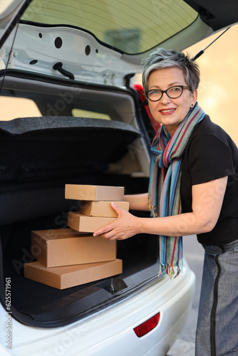Positive senior woman is loading cardboard boxes into car trunk.