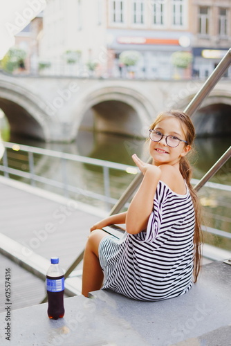 Portrait of little charming happy girl in glasses sits on bund of canal river in summer