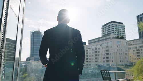 Rear back view following handsome male businessman in suit walks roof top terrace in office building overlooking modern city downtown cityscape with high-rise skyscrapers backlit with bright sunlight photo