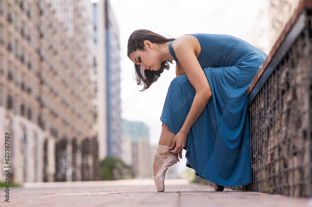Beautiful asian ballerina in blue dress adjusting her pointe shoes outdoors. 