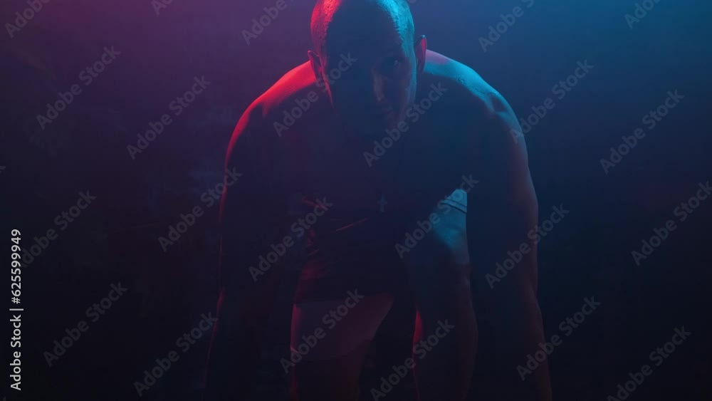 Male bodybuilder crouched down and getting ready to start, looking at the camera, in neon light in smoke in a dark room. Male bodybuilder crouched down and getting ready to start, looking at the camera, in neon light in smoke in a dark room.
