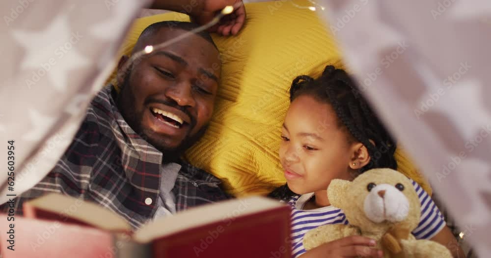 Happy african american father and daughter lying in tent and reading book