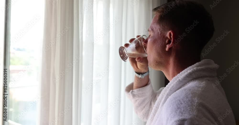 Satisfied young man in white bathrobe drinks cold refreshing water from glass standing in front of bright large window. Relaxing man looks out of window enjoying view