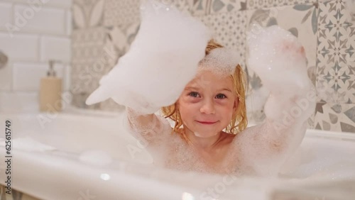 Cute caucasian blonde three years old girl taking bath playing with white soap foam bubbles,looking at camera.Carefree childhood, bathroom games concept.