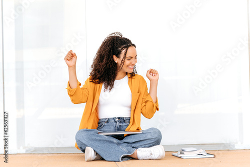 Success, win. Joyful curly haired brazilian or hispanic female student, sits on the floor near the university campus, finished studying online, rejoicing in luck, gesturing with both hands, smiling