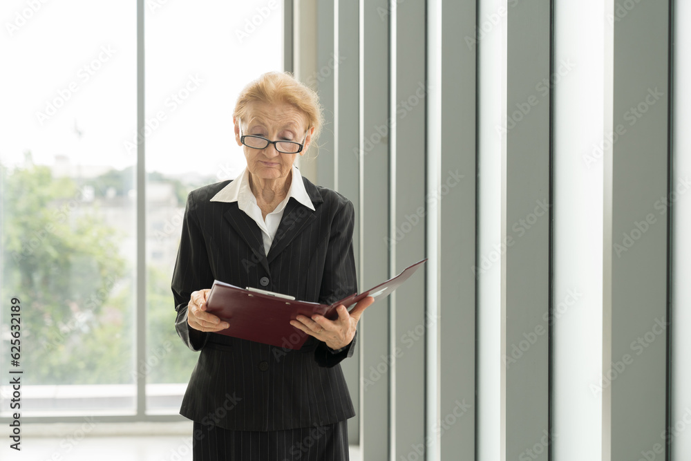 Senior business man and office. Elderly business woman working in office. Smiling older business woman standing and holding clipboard with blur background office Senior business man and office. Elderly business woman working in office. Smiling older business woman standing and holding clipboard with blur background office