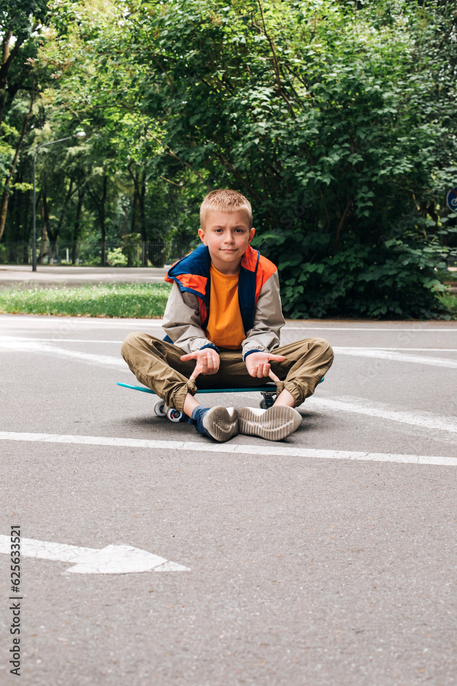 Outdoor activities for children.A cheerful young boy in bright clothes sits on a skateboard. Caucasian schoolboy on a walk. Front view Outdoor activities for children.A cheerful young boy in bright clothes sits on a skateboard. Caucasian schoolboy on a walk. Front view