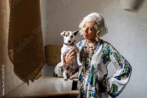 Senior woman standing in room and carrying pet dog in hand photo