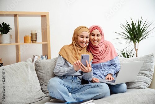 Portrait of beautiful happy arab girls in hijab working at home using smartphone and laptop while sitting on sofa. Women looking and typing on her mobile phone, holds a laptop