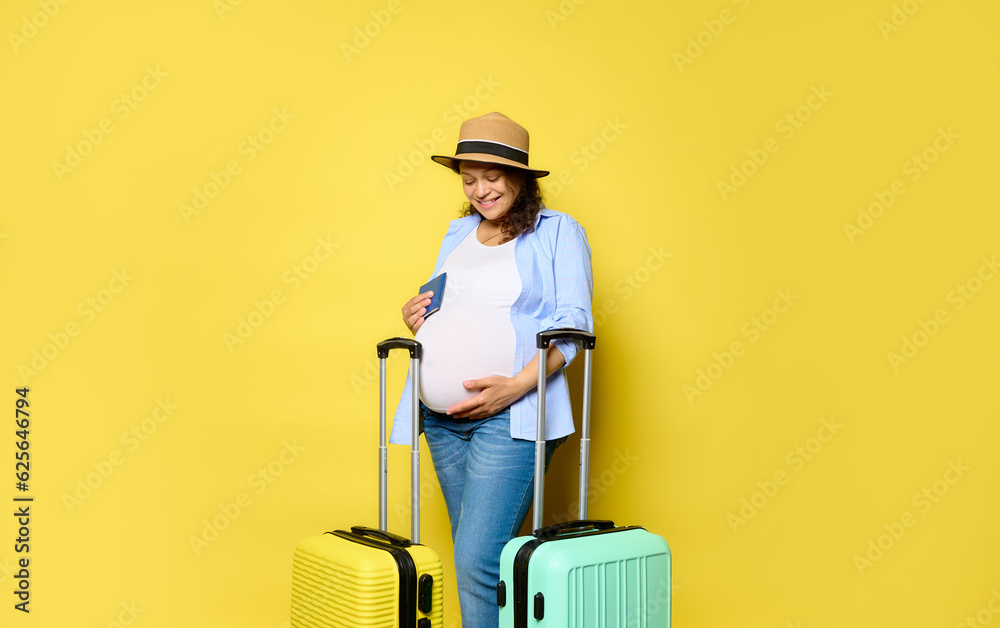 Smiling pregnant woman holding her belly, standing with boarding pass and suitcases, isolated yellow studio background Smiling pregnant woman holding her belly, standing with boarding pass and suitcases, isolated yellow studio background