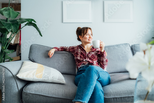 Young smiling woman sitting on sofa and looking side up while drinking coffee or tea. Young brunette woman relaxing after housekeeping, home cleaning. Portrait of relaxed female resting at home