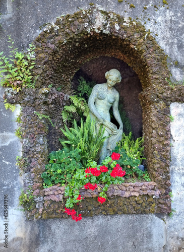 Romantic idyllic stone grotto in an antique style tropical garden with a delicate sculpture of  female statue and vibrant red flowers and green grass