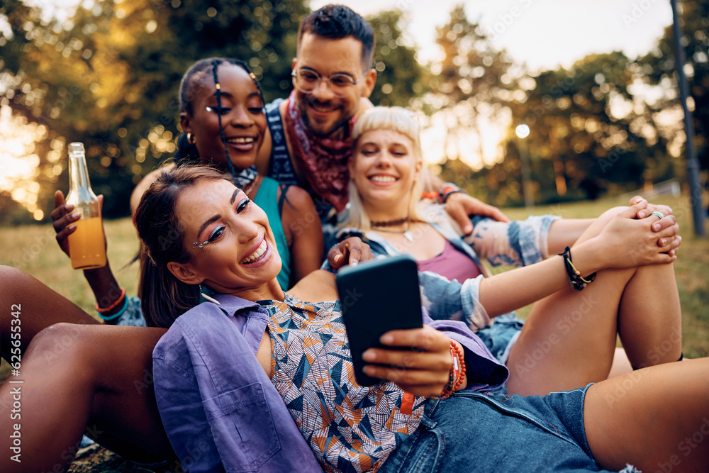 Fototapeta premium Happy woman taking selfie with her friends while relaxing on grass during summer music festival.