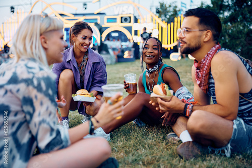 Multiracial group of happy festival goers eat burgers and drink beer while relaxing on grass.