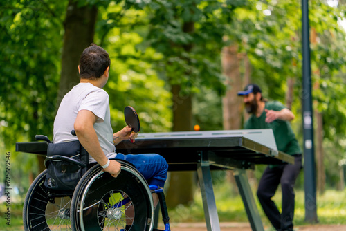 Inclusiveness A disabled man in a wheelchair plays ping pong with an older man in a city park against a backdrop of trees photo