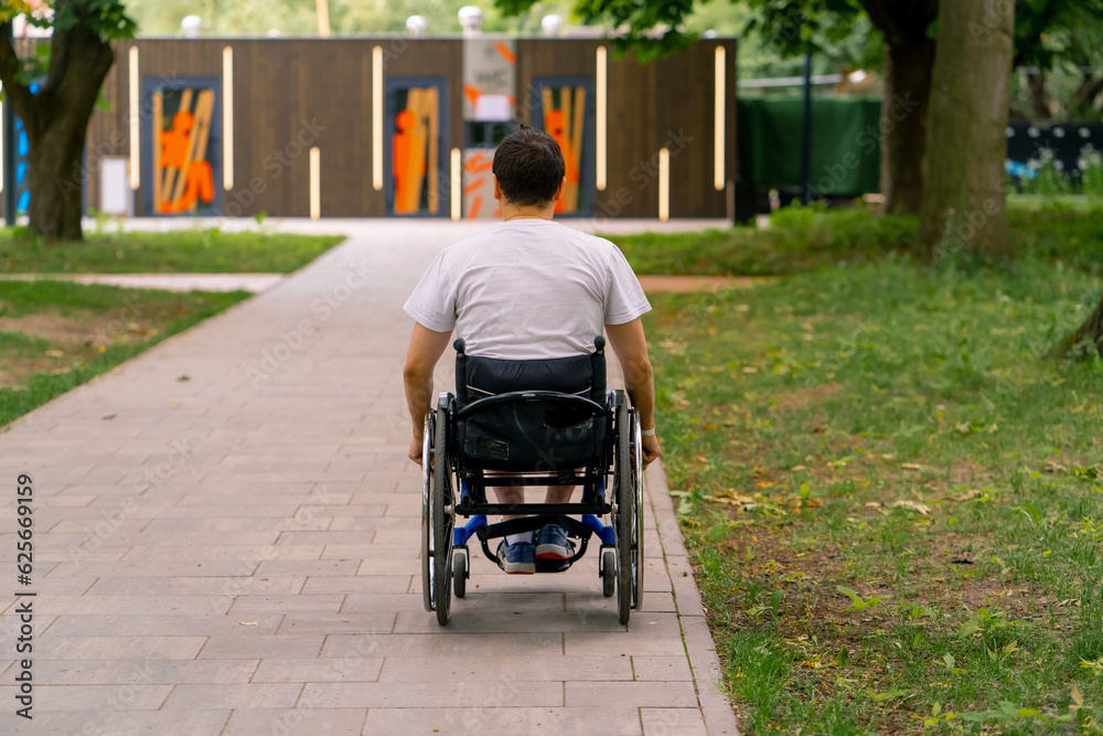 Obraz premium Inclusiveness A man with disabilities rides a wheelchair along the path of a city park against the background of trees Back view
