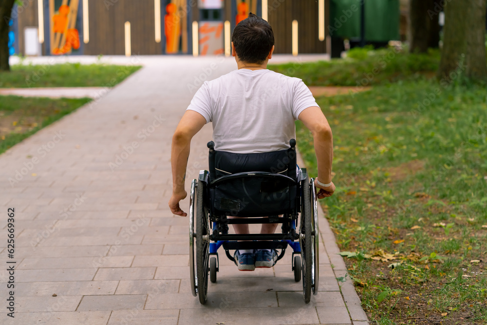 Obraz premium Inclusiveness A man with disabilities rides a wheelchair along the path of a city park against the background of trees Back view