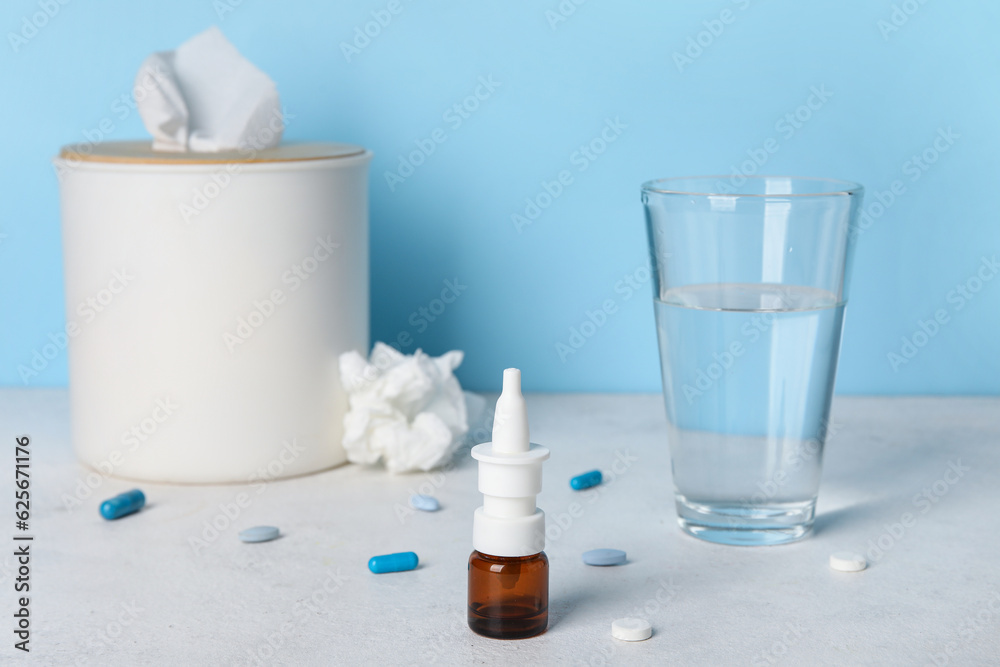 Nasal drops with pills, glass of water and tissue box on table near blue wall. Allergy concept