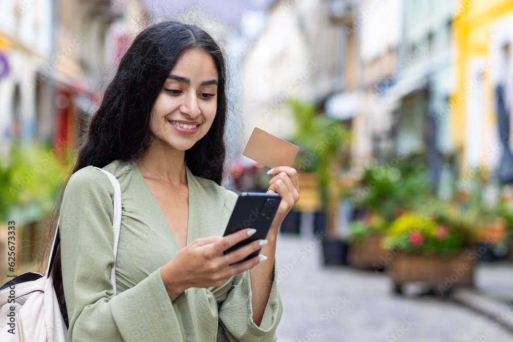 Young smiling Indian woman walking in the city, woman holding a bank credit card and phone, tourist making online booking of accommodation and booking tourist services while walking in the city