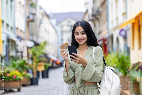 Young smiling Indian woman walking in the city, woman holding a bank credit card and phone, tourist making online booking of accommodation and booking tourist services while walking in the city photo