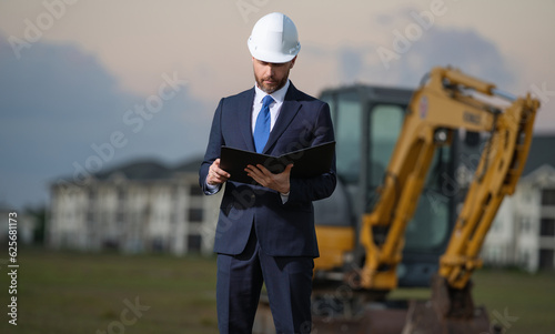 Construction owner near excavator. Confident construction owner in front of house. Architect, civil engineer. Man construction owner with a safety vest and hardhat at construction site.