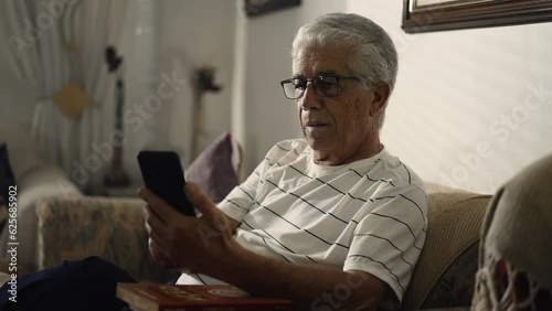 Elderly man staring at cellphone device sitting at home couch with concerned expression, watching video media online with baffled emotion