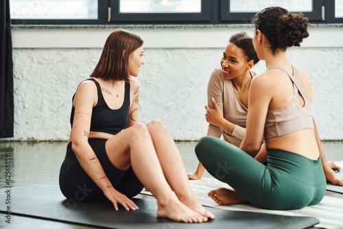 young and positive african american woman in sportswear sitting on yoga mat and talking to tattooed girlfriend during yoga class in gym, friendship, harmony and mental health concept photo