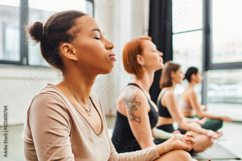 young african american woman meditating with closed eyes near multiethnic friends relaxing in easy pose during yoga class, harmony and wellness concept