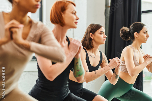 young and tattooed woman looking forward while practicing yoga with clenched hands near multiethnic girlfriends on blurred foreground, harmony and mental health concept
