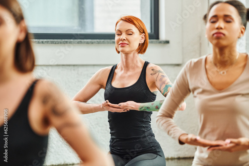 tattooed and redhead woman practicing yoga and meditating with closed eyes near multicultural women on blurred foreground, inner peace and body awareness concept © LIGHTFIELD STUDIOS