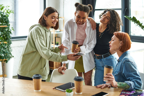 joyous young woman showing mobile phone to multicultural friends near takeaway drinks and magazines on table in interest club, happy gathering and leisure of diverse female team