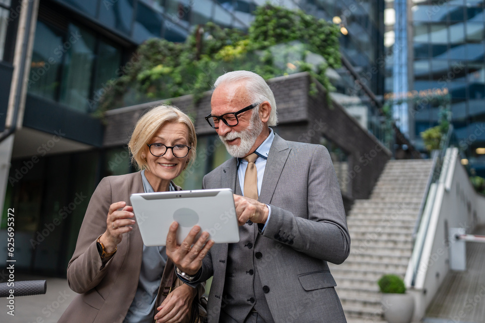 senior man dressed in a suit and a mature blonde woman as business partners husband and wife friends or colleagues talk With digital tablet in hand browse internet look data exchange ideas copy space senior man dressed in a suit and a mature blonde woman as business partners husband and wife friends or colleagues talk With digital tablet in hand browse internet look data exchange ideas copy space