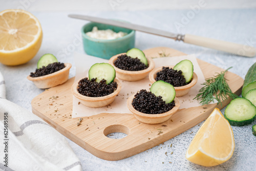 Wooden board of tasty tartlets with black caviar on grey background