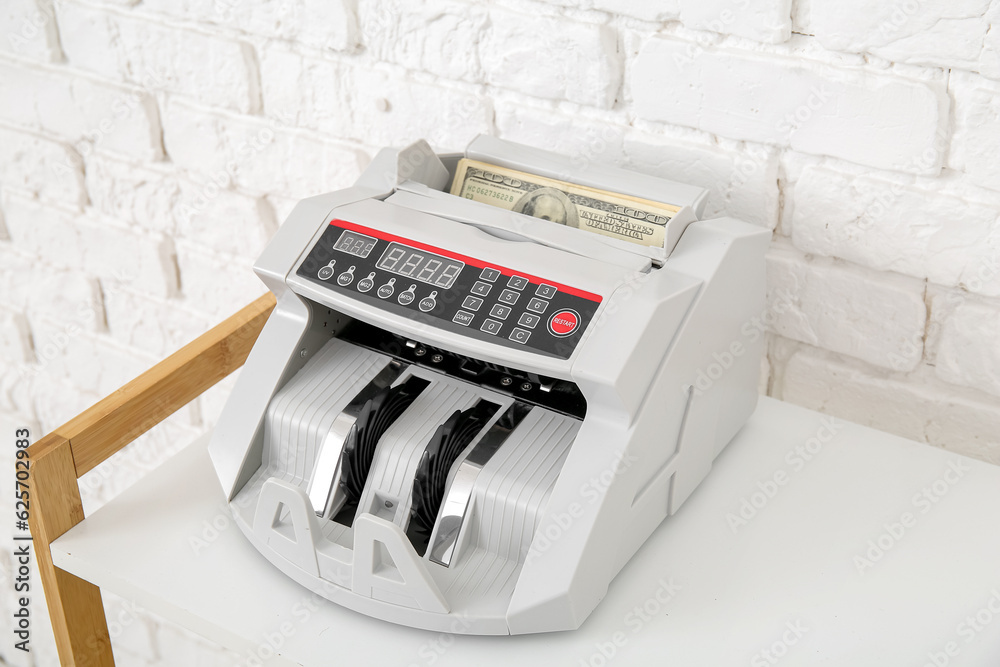 Cash counting machine with dollar banknotes on shelving unit near white brick wall, closeup