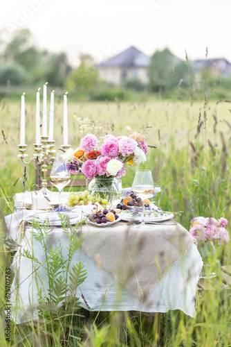 Dinner table for outdoor wedding or romantic date for a couple in a field. Elegant decoration with vintage chandelier, ranunculi flowers in vase, light snacks, cheese, fruits, white wine.