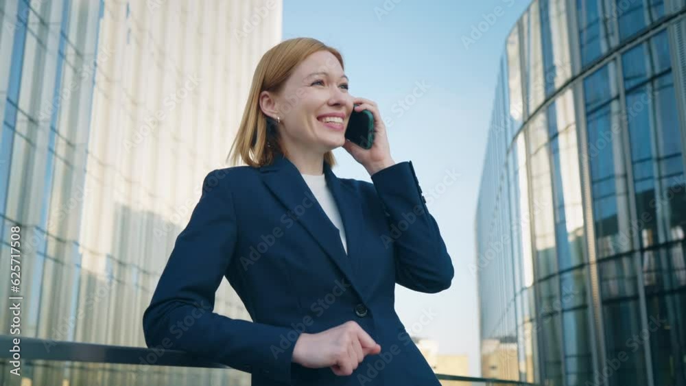 Gimbal shot positive blond hair business woman wearing blue formal suit talking on smartphone and smiles standing on office building rooftop leans on glass railing of terrace in city downtown