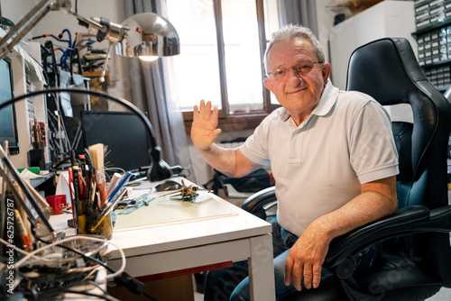 Portrait of smiling senior man wearing eyeglasses, waving hand, working in modern workshop at home