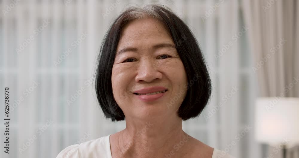 Closeup face portrait of happy and healthy Asian senior elderly gray and black-haired female smiling at camera. The old woman standing in living room with a smile. Happiness of elderly concept. Closeup face portrait of happy and healthy Asian senior elderly gray and black-haired female smiling at camera. The old woman standing in living room with a smile. Happiness of elderly concept.