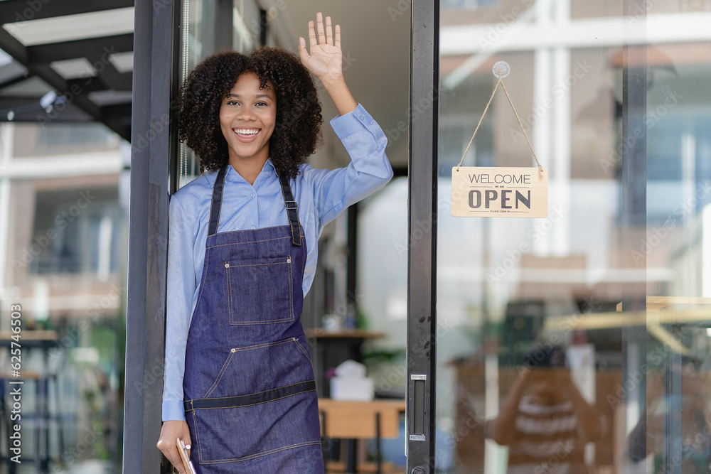 young african woman starting a small business working in a cafe standing in front of the store with open sign and greeting customers with happy smile, food and beverage business concept