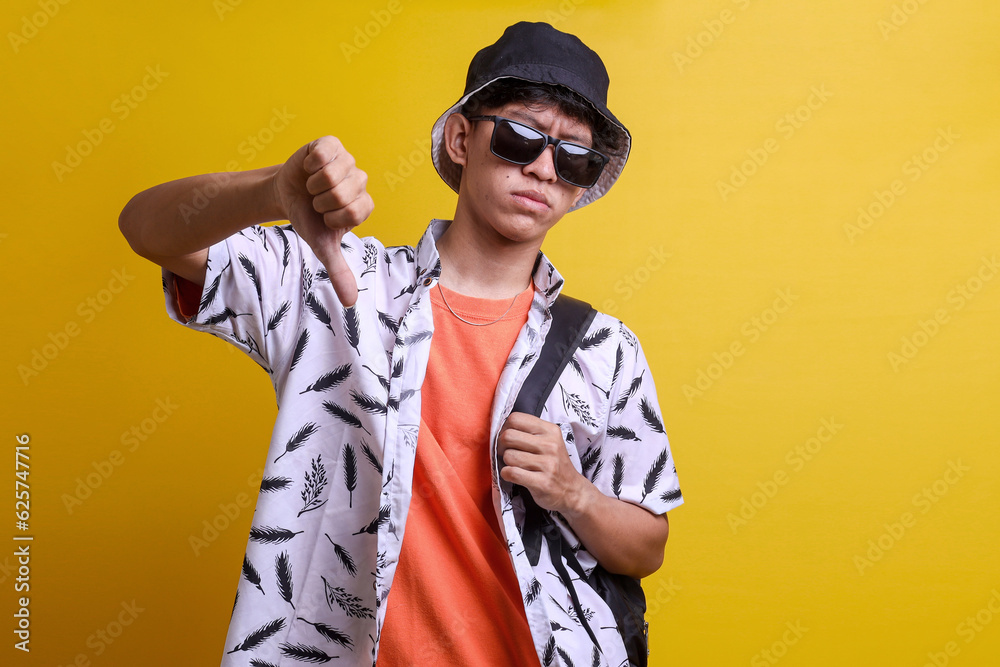Young unhappy dissatisfied man wear orange t-shirt, bucket hat and sunglasses, show thumb down dislike gesture isolated on yellow background. People lifestyle concept Young unhappy dissatisfied man wear orange t-shirt, bucket hat and sunglasses, show thumb down dislike gesture isolated on yellow background. People lifestyle concept