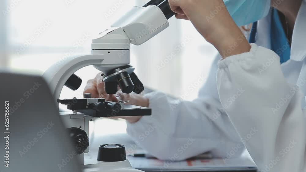 Medical worker in lab coat and sterile mask, doing a microscope analysis while her colleague are working behind